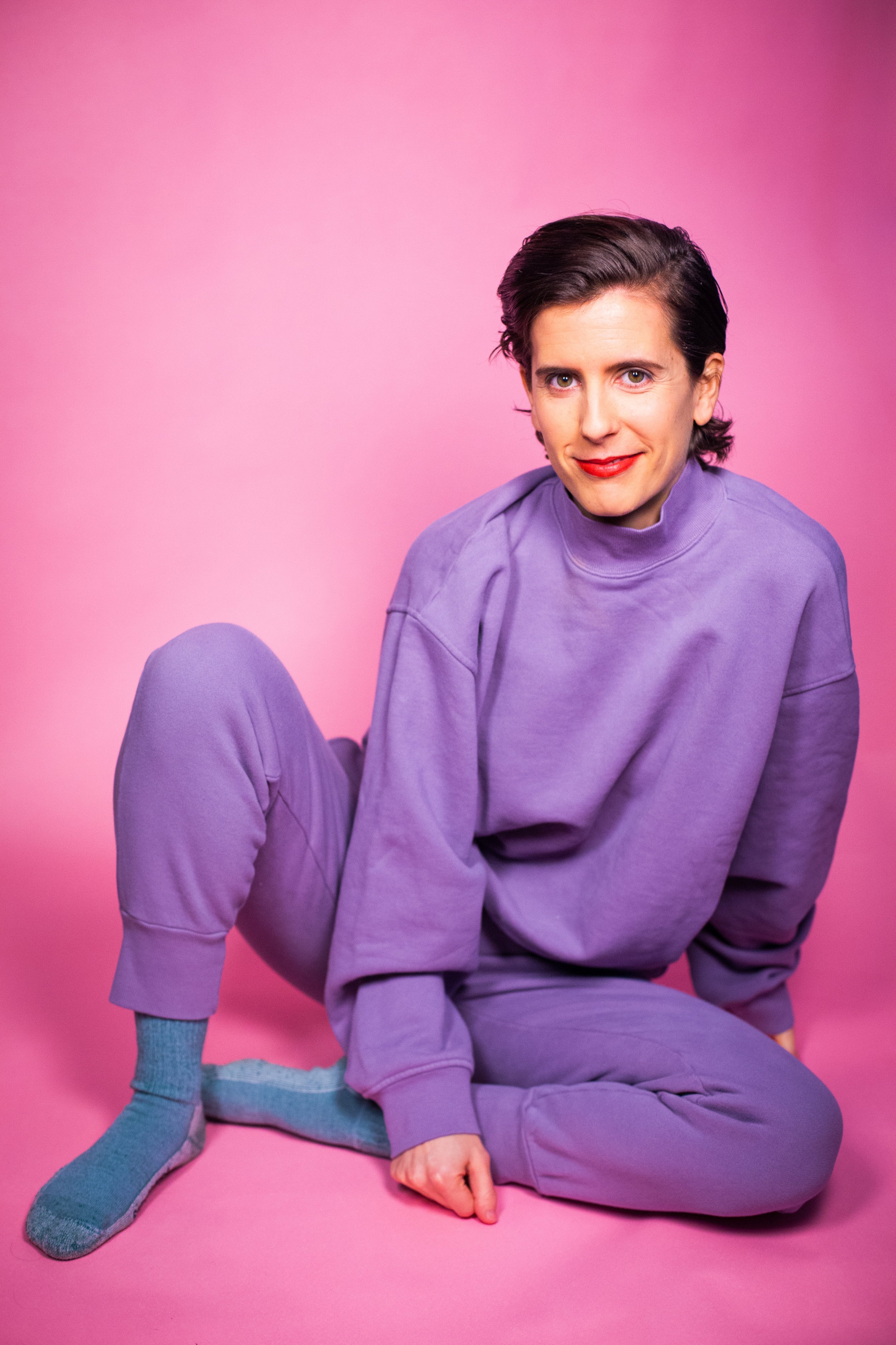 Portrait of KK Apple smiling directly at the camera, sitting on the ground in purple sweatsuit on a pink backdrop.